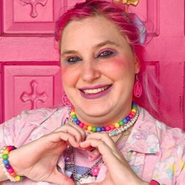 A photo of the artist. A pale skinned person with bright pink hair pulled up in a pony tail. purple, pink and blue bows decorate their hair. They have a wide smile, and vibrant blue and pink makeup. They also wear layers of vibrant necklaces. One is rainbo